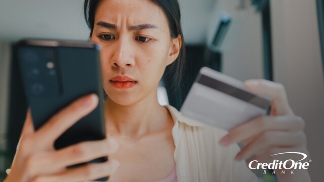 A woman holds her credit card while looking at her smartphone with a concerned expression, as if she was just notified of an unknown credit card charge.